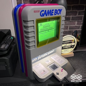 Könnte beinhalten: Ein grauer Nintendo Game Boy Arcade-Automat mit blauen, rosa und roten Akzenten. Der Bildschirm zeigt "Super Mario Land". Der Text "GAME BOY" ist blau. Der Automat hat einen Game Boy-Controller und den Text "with portable power!"