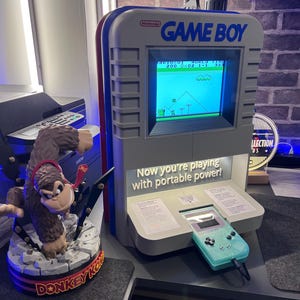 May include: A gray Game Boy arcade cabinet with red and blue accents, displaying a classic video game. A mint green Game Boy is connected. A Donkey Kong statue is on the left. The text reads "Now you're playing with portable power!"