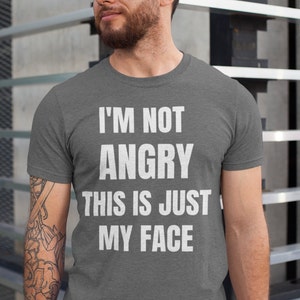 May include: A dark gray t-shirt with the text "I'm not angry this is just my face" printed in white.