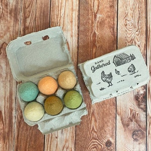 May include: An open egg carton with six colorful felt balls in shades of green, orange, and beige. A second egg carton is next to it, with the words "Hand Gathered" and a drawing of a barn and chickens.
