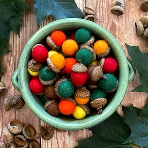 May include: A green ceramic bowl filled with colorful felt acorns. The acorns are in shades of red, orange, yellow, and green, with brown caps. The bowl sits on a wooden surface, with scattered acorns and green leaves.