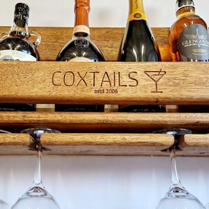 K&ouml;nnte beinhalten: Ein Wand-Weinregal aus Holz mit den Worten "COXTIALS estd 2006" und einem eingravierten Martini-Glas-Design auf der Vorderseite. Das Regal bietet Platz f&uuml;r sechs Flaschen Alkohol und hat zwei h&auml;ngende Weingl&auml;ser.
