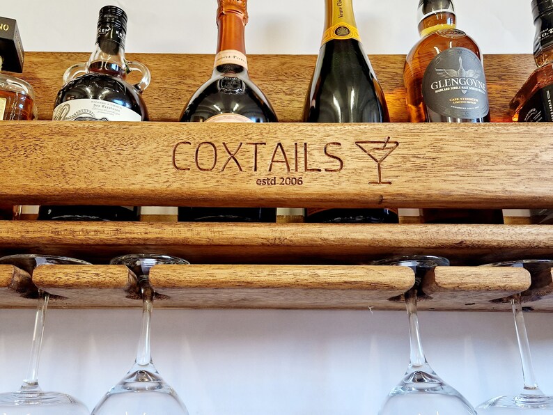 K&ouml;nnte beinhalten: Ein Wand-Weinregal aus Holz mit den Worten "COXTIALS estd 2006" und einem eingravierten Martini-Glas-Design auf der Vorderseite. Das Regal bietet Platz f&uuml;r sechs Flaschen Alkohol und hat zwei h&auml;ngende Weingl&auml;ser.