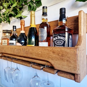 K&ouml;nnte beinhalten: Ein wandmontierter Weinst&auml;nder aus Holz mit sechs Gl&auml;sern, die darunter h&auml;ngen. Der St&auml;nder h&auml;lt f&uuml;nf Flaschen Alkohol, darunter Jack Daniel's Tennessee Whiskey, eine Flasche Wei&szlig;wein und eine Flasche Rotwein. Der St&auml;nder ist in einem dunklen Braunton gebeizt.