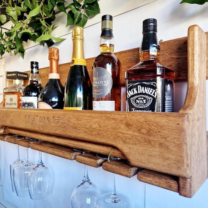K&ouml;nnte beinhalten: Ein wandmontierter Weinst&auml;nder aus Holz mit sechs Gl&auml;sern, die darunter h&auml;ngen. Der St&auml;nder h&auml;lt f&uuml;nf Flaschen Alkohol, darunter Jack Daniel's Tennessee Whiskey, eine Flasche Wei&szlig;wein und eine Flasche Rotwein. Der St&auml;nder ist in einem dunklen Braunton gebeizt.