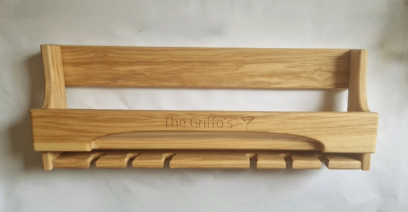 K&ouml;nnte beinhalten: Ein Wand-Weinglashalter aus Holz mit einer personalisierten Inschrift "The Griffo's" und einem Weinglas-Symbol. Der Halter hat ein Regal und eine Reihe von Haken zum Aufh&auml;ngen von Weingl&auml;sern.