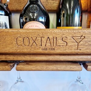 K&ouml;nnte beinhalten: Ein Wand-Weinregal aus Holz mit den Worten "COXTALS estd 2006" und einem gravierten Martini-Glas. Das Regal hat vier Glashalter und enth&auml;lt verschiedene Flaschen mit Spirituosen.