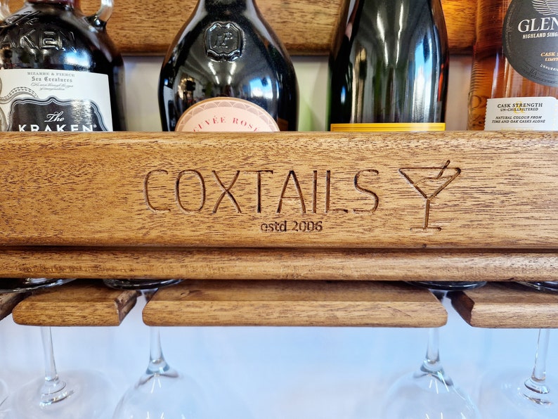 K&ouml;nnte beinhalten: Ein Wand-Weinregal aus Holz mit den Worten "COXTALS estd 2006" und einem gravierten Martini-Glas. Das Regal hat vier Glashalter und enth&auml;lt verschiedene Flaschen mit Spirituosen.