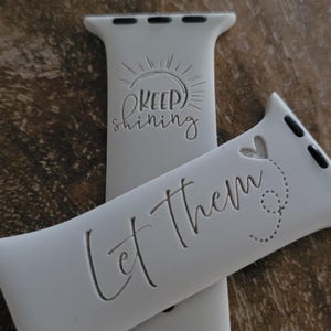 May include: White Apple Watch band with two different engraved messages. The top band says "Keep Shining" with a sun graphic. The bottom band says "Let Them" with a heart and a dotted line.