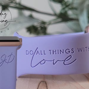 Könnte beinhalten: Ein lavendelfarbenes Silikonarmband für die Apple Watch mit dem eingravierten Text "Do all things with love". Das Armband ist an einer roségoldenen Apple Watch zu sehen.