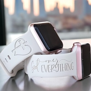 Może przedstawiać: Dwa białe paski do Apple Watch z różnymi wzorami. Jeden pasek ma serce z krzyżem w środku, a drugi pasek ma napis "Bóg ponad wszystko" kursywą.
