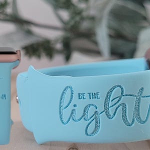 Puede incluir: Una banda de reloj Apple de silicona azul claro con las palabras "Sé la luz" y la escritura "Mateo 5:14" grabadas en ella.