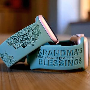 May include: Two teal Apple Watch bands. One band features a detailed black mandala design. The other band is engraved with the words "GRANDMA'S BLESSINGS" and the names Weller, Ariana, River, and Anakin. The watch hardware is rose gold.