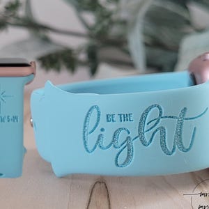 Puede incluir: Una banda de reloj de silicona azul claro con las palabras "Sé la luz" grabadas en ella. La banda es compatible con Apple Watch y tiene un acento de oro rosa.