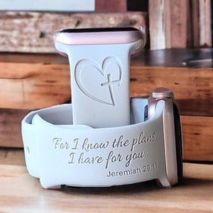 May include: White silicone Apple Watch band with a heart and cross design. The band has the text "For I know the plans I have for you... Jeremiah 29:11" engraved on it.