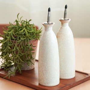 Può includere: Due bottiglie di olio e aceto in ceramica bianca con tappi argentati e beccucci neri su un vassoio di legno. Una pianta verde in vaso è sullo sfondo.