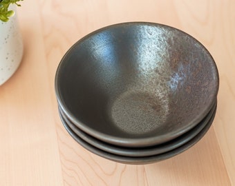 May include: Three black ceramic bowls stacked on top of each other. The bowls have a speckled glaze and a slightly iridescent finish.