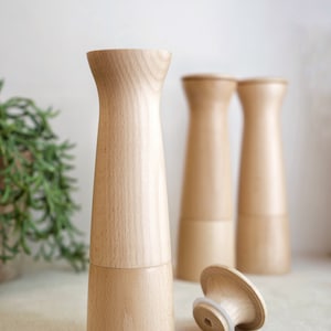 May include: Three wooden salt and pepper shakers with a natural wood finish. The shakers are cylindrical with a slightly flared top and bottom. One shaker is lying on its side with the top off, revealing a white rubber seal.
