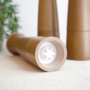 May include: Close-up of a wooden salt and pepper grinder set. The grinder on the left has a white plastic mechanism visible. The grinders are made of light brown wood.