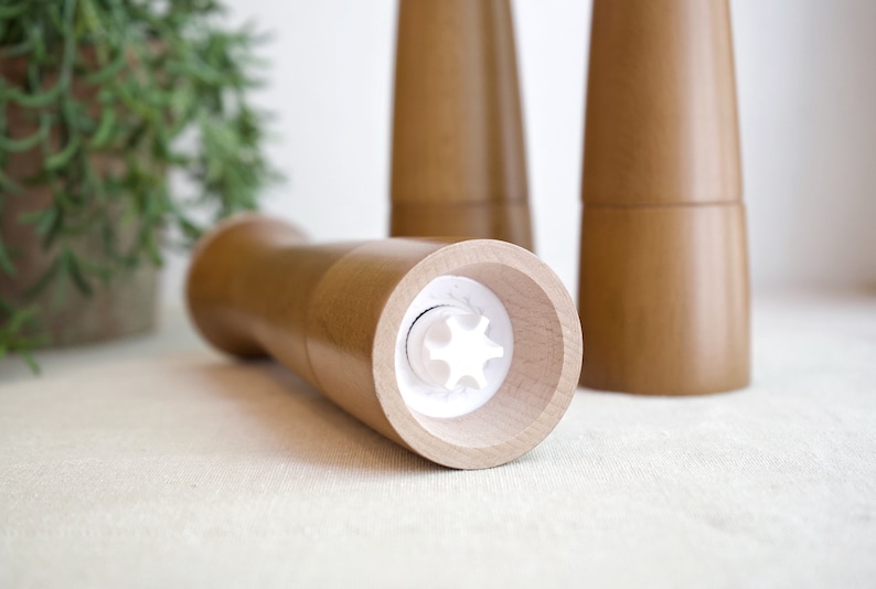 May include: Close-up of a wooden salt and pepper grinder set. The grinder on the left has a white plastic mechanism visible. The grinders are made of light brown wood.