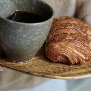 May include: A wooden tray with a cup of coffee and a croissant. The tray is being held by a person's hands.