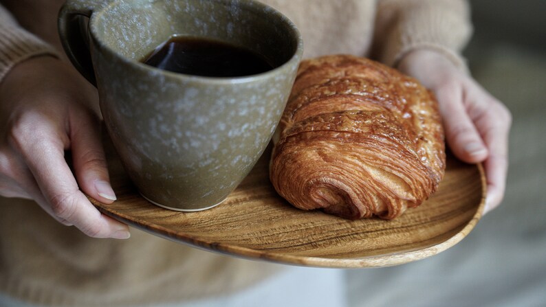 May include: A wooden tray with a cup of coffee and a croissant. The tray is being held by a person's hands.