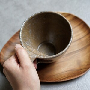 May include: A brown ceramic mug with a speckled glaze sits on a wooden coaster. The mug has a rounded handle.