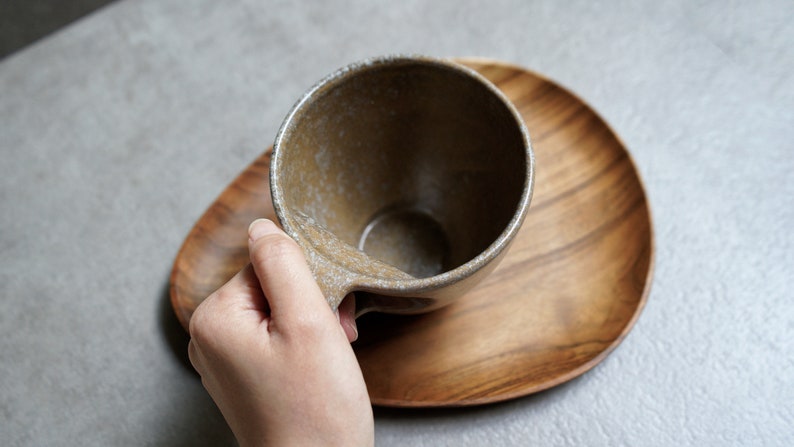 May include: A brown ceramic mug with a speckled glaze sits on a wooden coaster. The mug has a rounded handle.