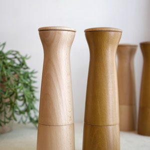 May include: Two wooden salt and pepper shakers, one light brown and one dark brown, with a simple design. The shakers are on a white surface.