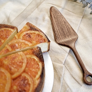 May include: A sliced orange cake with a wooden cake server on a neutral-coloured linen cloth. The cake is topped with orange slices. The wooden server has a long handle and a rounded end with a hole.