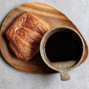 May include: A close-up of a wooden tray with a flaky pastry and a cup of black coffee. The coffee cup is a light brown colour with a speckled glaze.