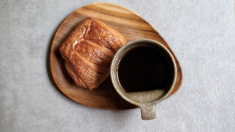 May include: A close-up of a wooden tray with a flaky pastry and a cup of black coffee. The coffee cup is a light brown colour with a speckled glaze.