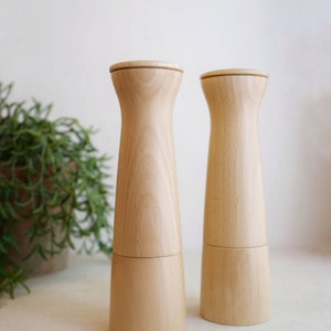 May include: Two wooden salt and pepper shakers with a light brown finish. The shakers are cylindrical with a slightly tapered top and bottom.