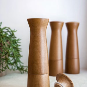 May include: Three wooden salt and pepper shakers with a natural wood finish. The shakers are cylindrical with a slightly flared top and bottom. One shaker is lying on its side, revealing a white rubber ring around the top.