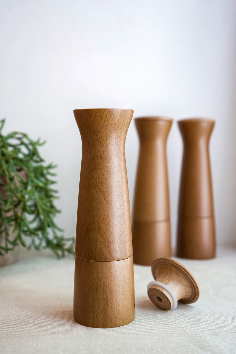 May include: Three wooden salt and pepper shakers with a natural wood finish. The shakers are cylindrical with a slightly flared top and bottom. One shaker is lying on its side, revealing a white rubber ring around the top.