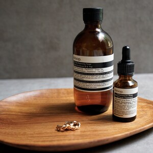 May include: Two amber glass bottles with black labels and white text on a wooden tray. The larger bottle is labelled "Aesop.  100 ml  Facial Toner" and the smaller bottle is labelled "Aesop.  Damascan Rose Facial Treatment". A gold ring is on the tray.