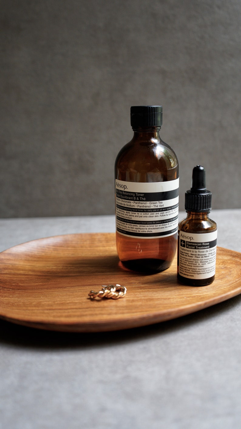 May include: Two amber glass bottles with black labels and white text on a wooden tray. The larger bottle is labelled "Aesop.  100 ml  Facial Toner" and the smaller bottle is labelled "Aesop.  Damascan Rose Facial Treatment". A gold ring is on the tray.