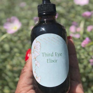 May include: A dark brown glass bottle with a black dropper top. The bottle has a light blue label with the words "Third Eye Elixir" and a floral design. The background is blurred with green foliage and pink flowers.