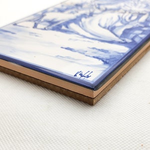 May include: A blue and white ceramic tile with a painted scene of a landscape. The tile has a brown backing and is signed "1444" in the bottom right corner.