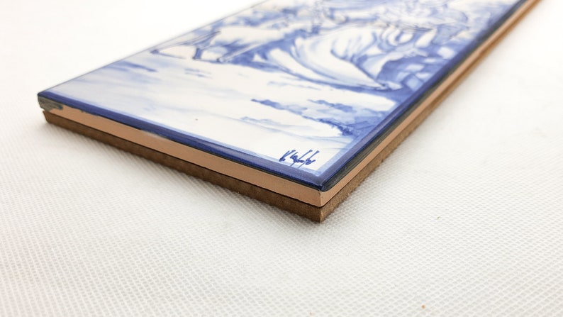 May include: A blue and white ceramic tile with a painted scene of a landscape. The tile has a brown backing and is signed "1444" in the bottom right corner.