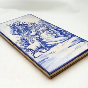 May include: A blue and white ceramic tile depicting a traditional nativity scene. The figures of Mary, Joseph, and the baby Jesus are painted in blue and white, with a lamb at their feet.