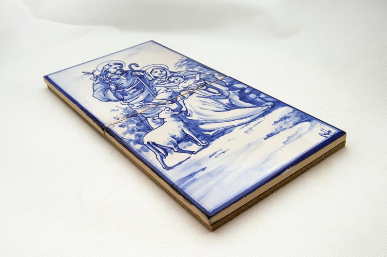 May include: A blue and white ceramic tile depicting a traditional nativity scene. The figures of Mary, Joseph, and the baby Jesus are painted in blue and white, with a lamb at their feet.