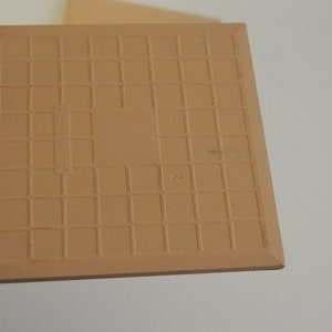 May include: A tan ceramic tile with a grid pattern on the back. The tile has a number "2" written on it.