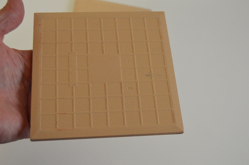 May include: A tan ceramic tile with a grid pattern on the back. The tile has a number "2" written on it.