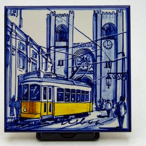 May include: A blue and white ceramic tile with a painted image of a yellow tram in front of a church with two towers. The tram is on a street with people walking. The tile has the word "Lisboa" written on it.