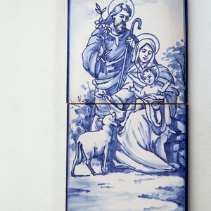 May include: A blue and white ceramic tile depicting the Holy Family. Joseph is holding a staff and a branch with leaves. Mary is holding the baby Jesus. A white lamb is in the foreground.