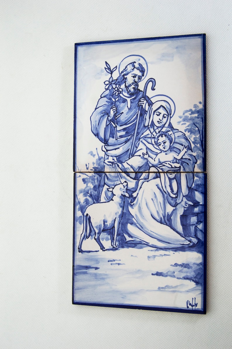 May include: A blue and white ceramic tile depicting the Holy Family. Joseph is holding a staff and a branch with leaves. Mary is holding the baby Jesus. A white lamb is in the foreground.