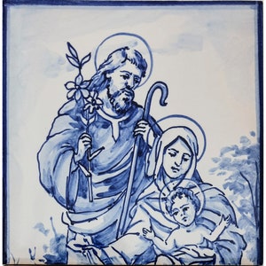 May include: A blue and white ceramic tile depicting a traditional depiction of the Holy Family. Joseph is on the left, holding a staff and a branch with flowers. Mary is on the right, holding baby Jesus. The tile has a blue and white background.