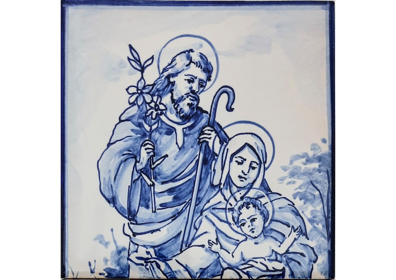 May include: A blue and white ceramic tile depicting a traditional depiction of the Holy Family. Joseph is on the left, holding a staff and a branch with flowers. Mary is on the right, holding baby Jesus. The tile has a blue and white background.