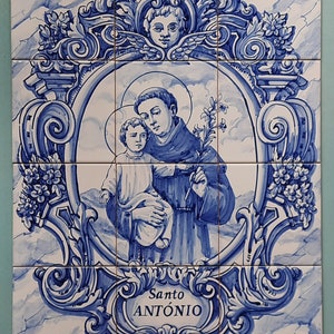 Könnte beinhalten: Eine blaue und weiße Keramikfliese, die den Heiligen Antonius von Padua zeigt, der ein Jesuskind hält. Die Fliese ist mit verzierten blauen und weißen Blumenmustern gerahmt und die Worte "Santo ANTONIO" sind auf einem blauen Banner unten geschrieben.
